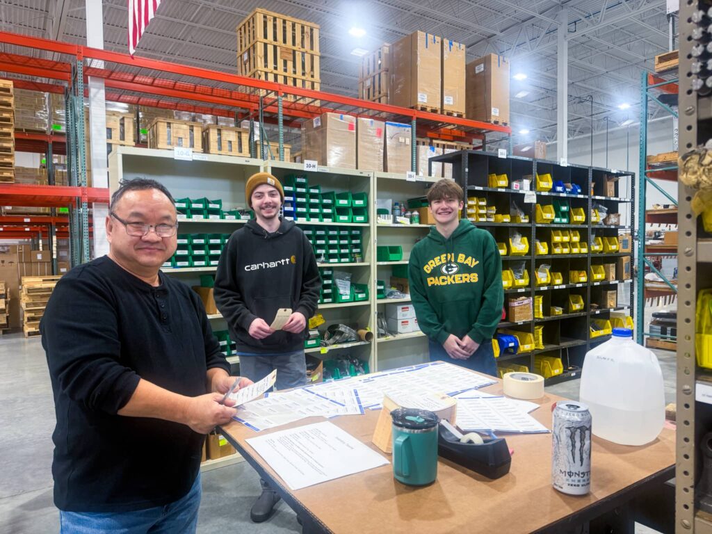 The photo shows three people working in a warehouse setting. The individuals are standing around a table with product labels and packing materials. The person on the left is holding a label, and the two others are standing next to him, smiling. The background features shelves stocked with boxes and parts. One person is wearing a Green Bay Packers hoodie, another has a Carhartt hoodie, and the third is holding a can of Monster energy drink.