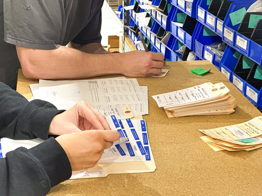 The photo shows two people working at a table, with their hands visible as they organize or label paperwork. There are several stacks of forms and labels on the table, some of which are being handled by the individuals. Behind them, there are neatly organized bins containing parts or supplies, each labeled with a number. The workspace has a clean and organized appearance, suggesting a task involving inventory management or labeling.