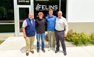 The photo shows four people standing in front of a building with a "Felins" logo on the window. The group consists of two men in the front and two men behind them. The person on the left is wearing a blue vest, beige pants, and brown shoes. The person next to him is wearing a black shirt, blue jeans, and sneakers. The man on the right is in a light blue shirt, gray pants, and dark shoes, while the last person is wearing a white shirt, dark pants, and black shoes. There are flowers and plants in the foreground, and the building’s window shows the "Felins" logo along with "Suite 400."