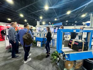 The image shows several people at a trade show booth, where they are engaging with a live demonstration of an automated packaging system. The machine, with blue and silver components, is bundling boxes together, and the boxes are marked with the "DURANGO" logo. One person is standing at the machine, while others are observing and discussing. The booth includes a sign in the background promoting sustainable packaging. There are plants near the machine, and other booths are visible in the background, indicating a busy and active trade show environment.