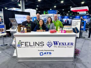 The image shows a group of five people standing at a booth at a trade show. The booth is branded with the Felins logo and the tagline “Simplify, Solve, Sustain.” The people, dressed in various company-branded attire, are smiling and posing behind the booth’s counter. The booth displays packaging materials such as rolls of tape, packaging products, and promotional items. In the background, other booths and attendees are visible, indicating a busy trade show environment. The booth is also proudly representing ATS Unique Banding, in partnership with Wexler.