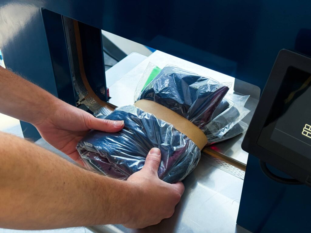 Close-up of a person placing bundled, poly-bagged garments into a Felins US-3000 banding machine using sustainable paper banding.