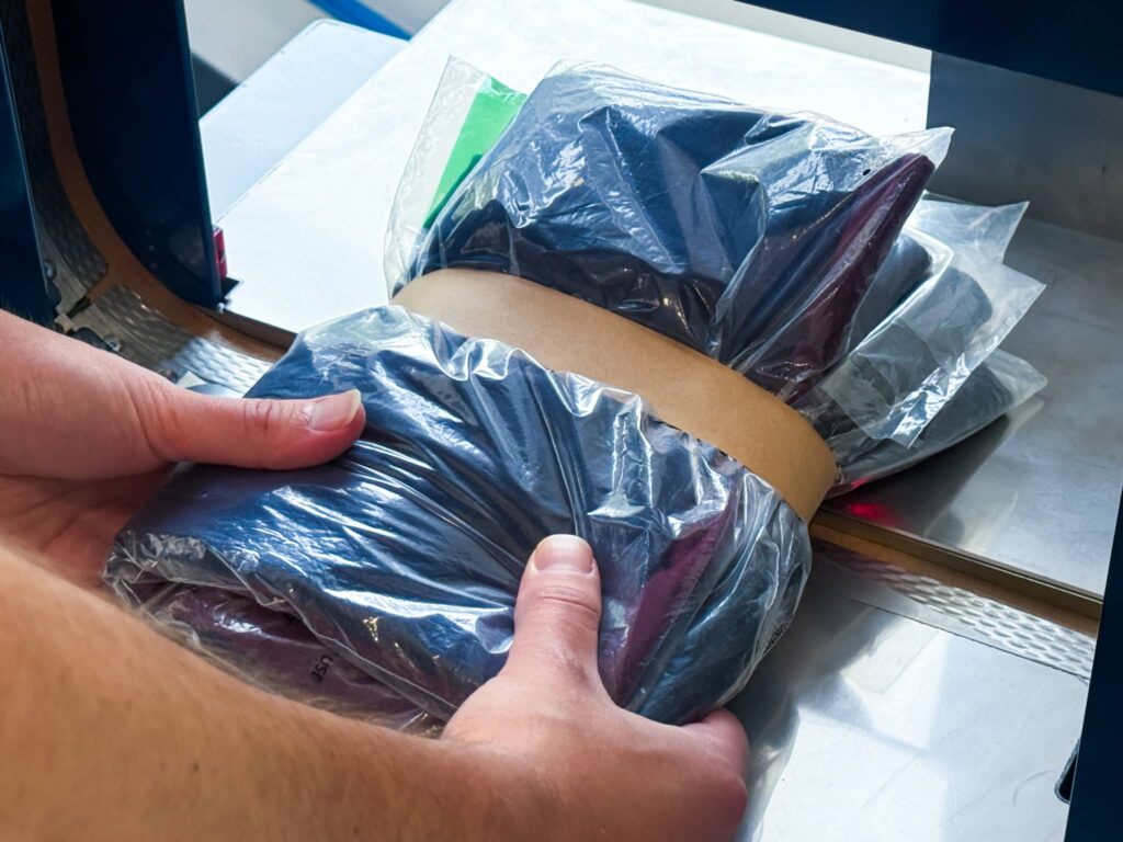 Hands holding a stack of plastic-wrapped apparel being secured with a recyclable paper band in the Felins US-3000 banding machine.
